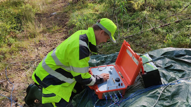 Henkilö käyttää oranssia mittalaitetta metsässä.