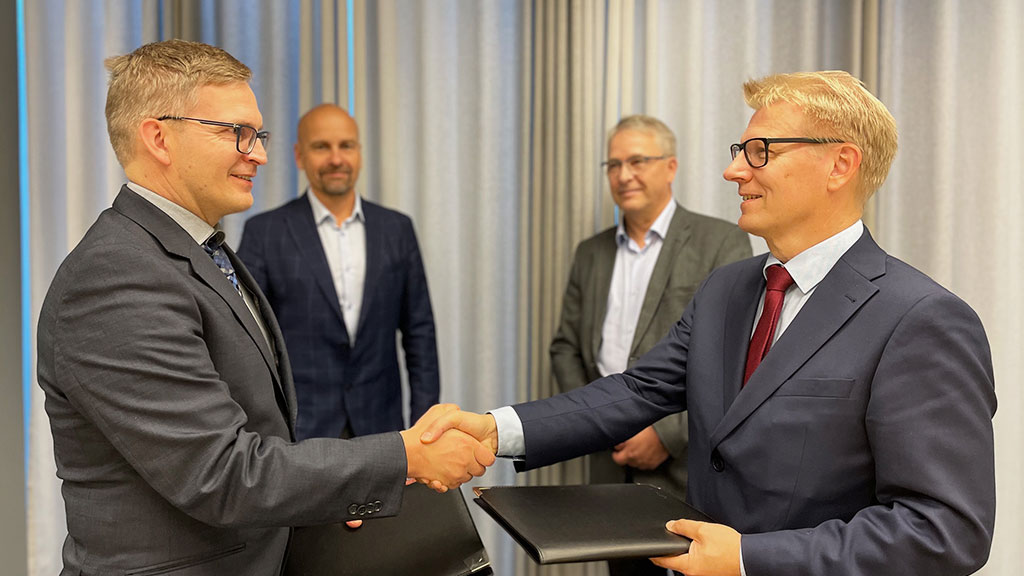 Sopimuksen allekirjoittaneet henkilöt kättelevät ja taustalla kaksi muuta miestä katsoo.