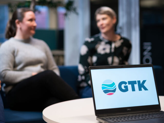 Etualalla läppäri GTK:n logolla, takana sohvalla istuvat ihmiset keskustelemassa