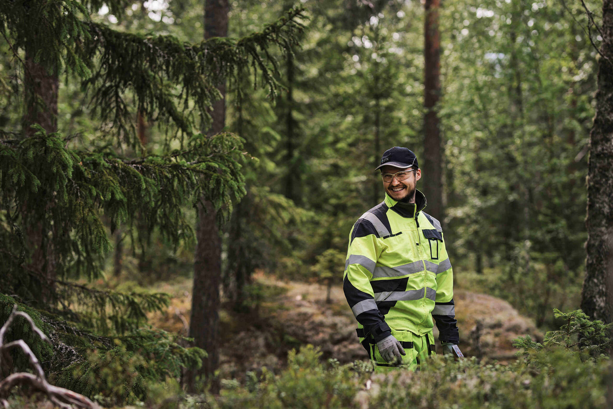 Hymyilevä mies huomiovaatteissa ja laseissa metsässä