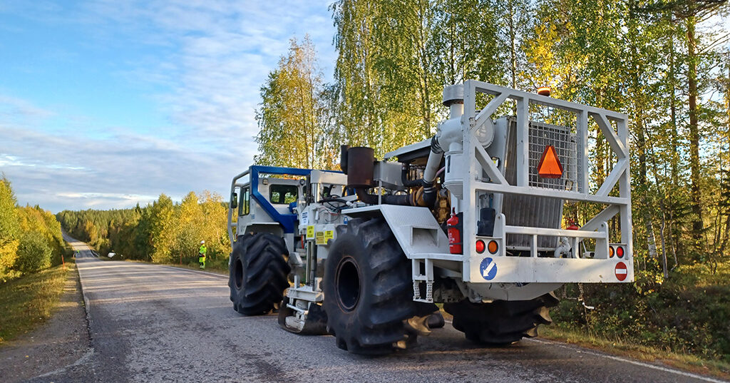 Tekninen, isopyöräinen ajoneuvo seisoo hiekkatiellä, jonka varrella kasvaa lehtipuita.