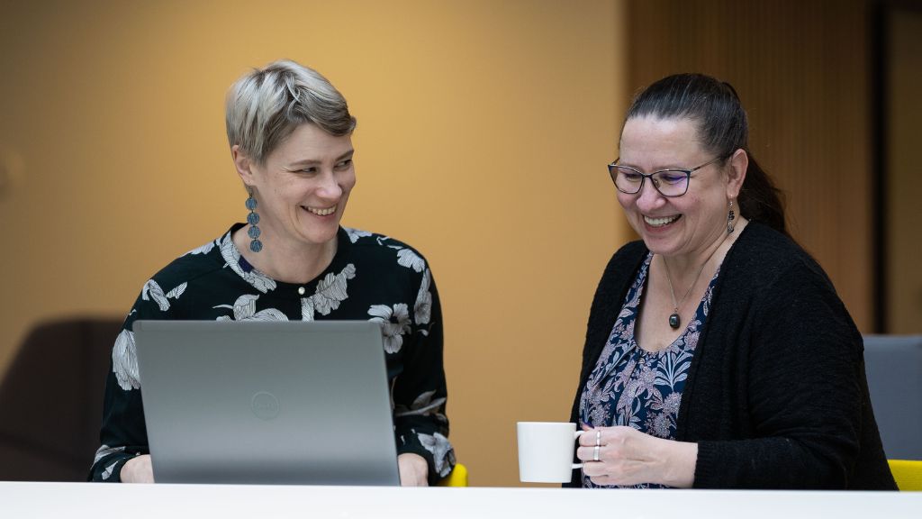 Two smiling persons sitting next to each other with a laptop and a mug.