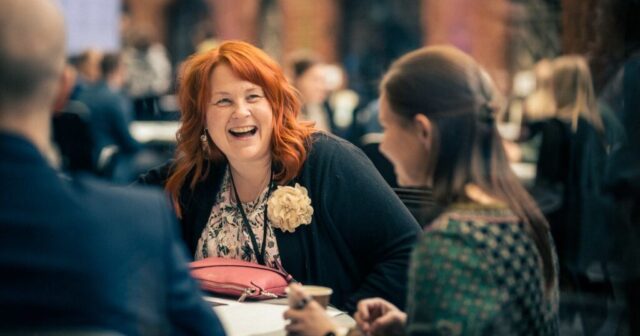 Red-haired woman sits by a table and laughs.