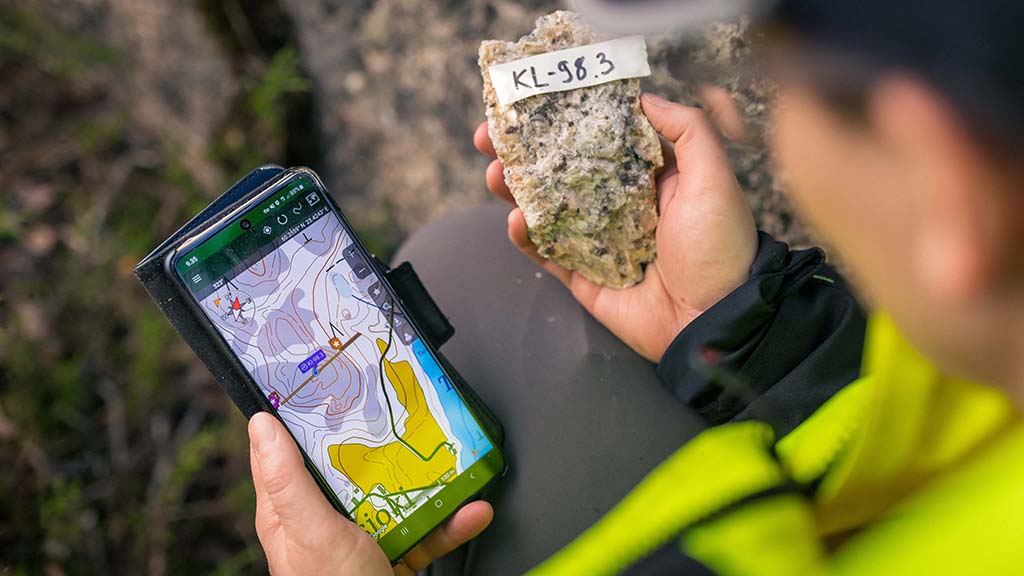 A geologist examines a rock sample in the field and uses a mobile device with a map application to document the sample location.