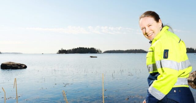 A woman dressed in a yellow work jacket looks over her shoulder at the camera, with a seascape in the background.