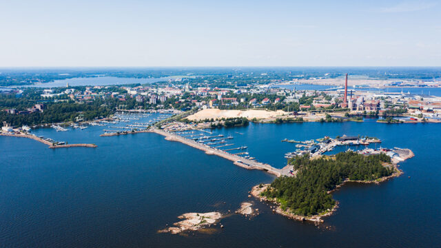 Ilmakuva Kotkan kaupungista mereltä päin. Merta, rannikkoa ja venesatama aurinkoisena kesäpäivänä. Kauempana taustalla Sunilan kaupunginosa.