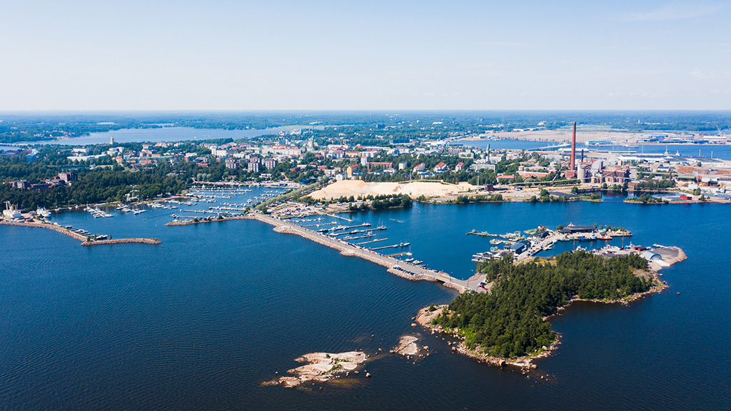 Ilmakuva Kotkan kaupungista mereltä päin. Merta, rannikkoa ja venesatama aurinkoisena kesäpäivänä. Kauempana taustalla Sunilan kaupunginosa.