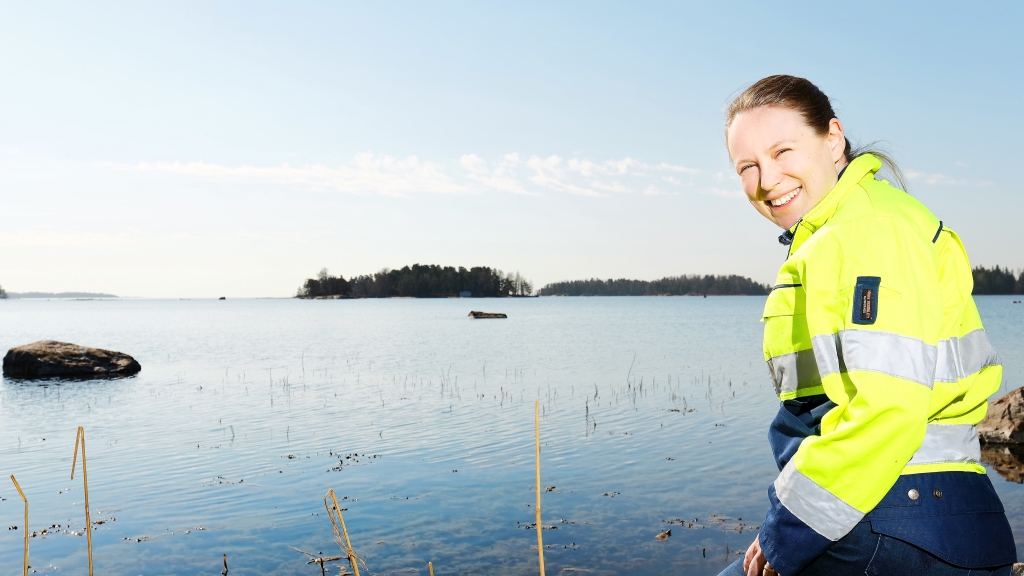 Keltaiseen työtakkiin pukeutunut nainen katsoo olkansa yli kameraan, taustalla merimaisema.