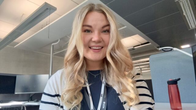 Hymyilevä henkilö istuu toimistossa työpisteen ääressä. Taustalla on tietokonenäyttöjä ja punainen juomapullo pöydällä.