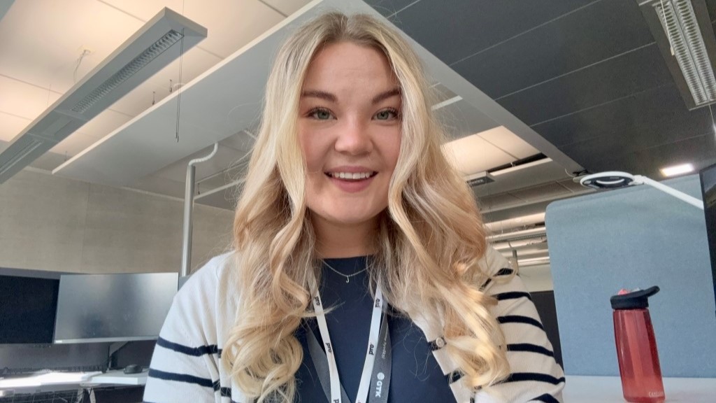Hymyilevä henkilö istuu toimistossa työpisteen ääressä. Taustalla on tietokonenäyttöjä ja punainen juomapullo pöydällä.