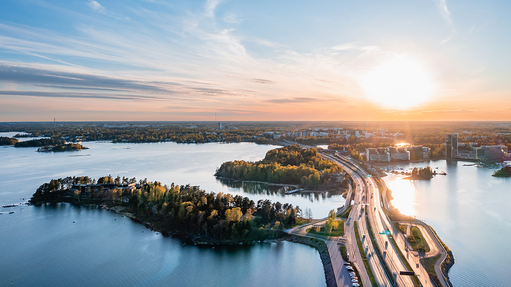 Näkymä ilmasta Helsingin Länsiväylälle auringon laskiessa. Merta ja vihreää luontoa, kaupunki taustalla.