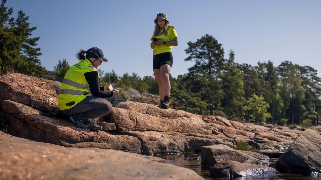 Kaksi ihmistä keltaisissa huomioliiveissä kivisellä rannalla. Toinen istuu ja kirjoittaa muistikirjaan, toinen seisoo ja katsoo puhelinta.