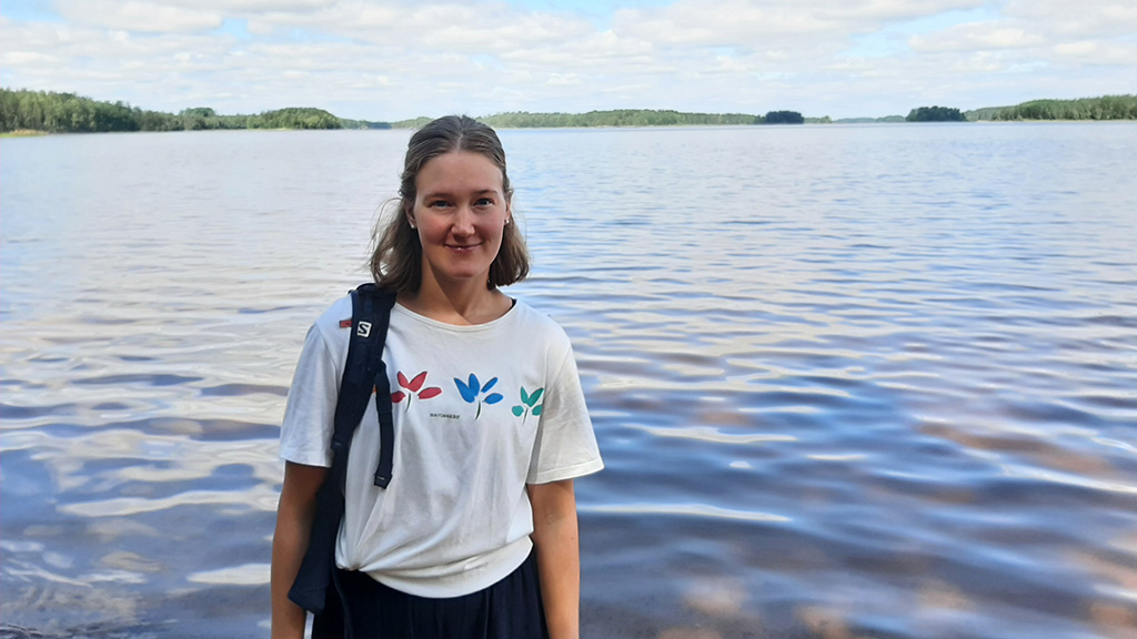 Hymyilevä henkilö kesäisessä asussa. Taustalla järvimaisema, jonka horisontissa näkyy saaria ja vihreää metsää.