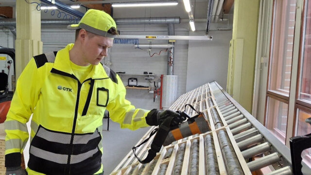 Keltaiseen työasuun pukeutunut henkilö tutkii kädessään olevalla laitteella kivilaatikoita.