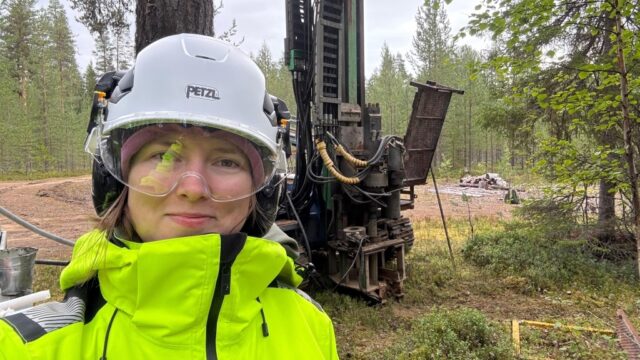 Henkilö käyttää suojakypärää ja suojalaseja metsässä porauslaitteen vieressä.