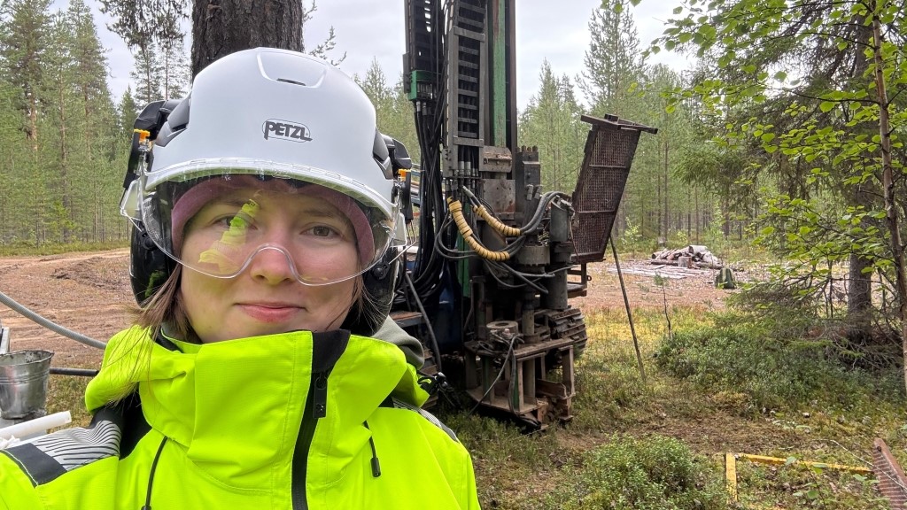 Henkilö käyttää suojakypärää ja suojalaseja metsässä porauslaitteen vieressä.
