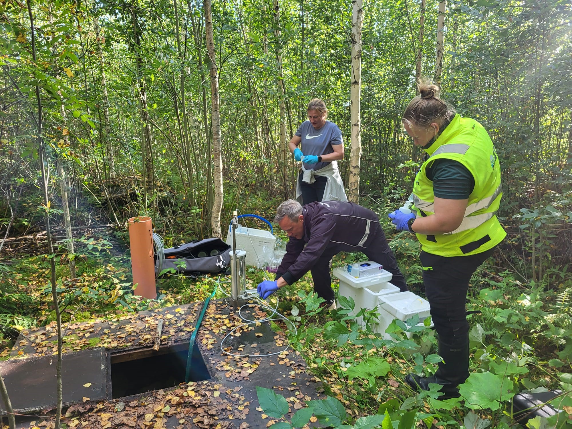 Kolme henkilöä ottaa vesinäytettä metsässä. Yksi henkilö on kumartunut luukun ääreen. Maassa on laitteita ja näyteastioita.
