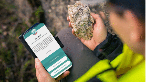 Henkilö pitelee kiveä toisessa kädessä ja puhelinta toisessa kädessä, näytöllä Omakivi-sovellus.