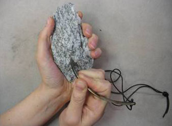 Käsi pitää kivinäytettä ja toisen käden koukkutyökalu raapii sen pintaa.