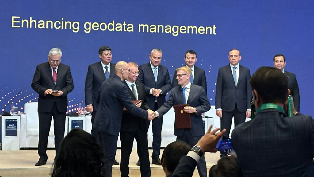 Ryhmä henkilöitä seisoo lavalla taustalla, jossa lukee "Enhancing geodata management". Kaksi henkilöä kättelee etualalla, ja ihmiset ottavat kuvia.