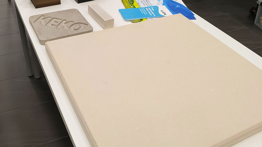 Valkoisella pöydällä on vaaleita betonilaattoja, joista yhdessä lukee "KEKO", sekä sinisiä esitteitä.