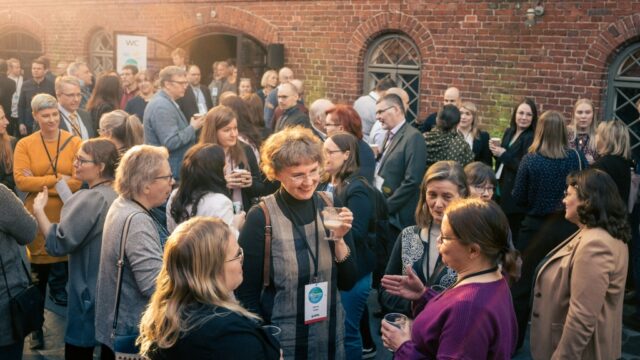 Suuri joukko ihmisiä seisoo ulkona tiiliseinän edessä, keskustelee ja pitelee juomia.