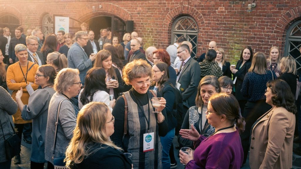 Suuri joukko ihmisiä seisoo ulkona tiiliseinän edessä, keskustelee ja pitelee juomia.