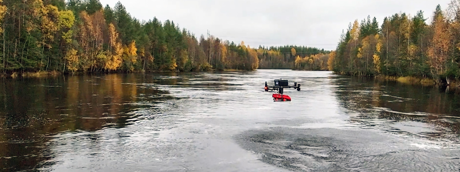 Jokea ympäröi syksyinen metsä, ja veden pinnalla kulkee tutkimuslaite tai drone, jota käytetään vesistöjen mittaukseen tai seurantaan.