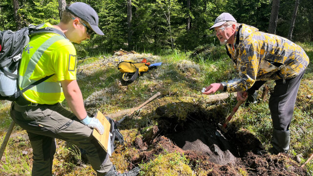 Two people dig soil in a forest next to a hole.