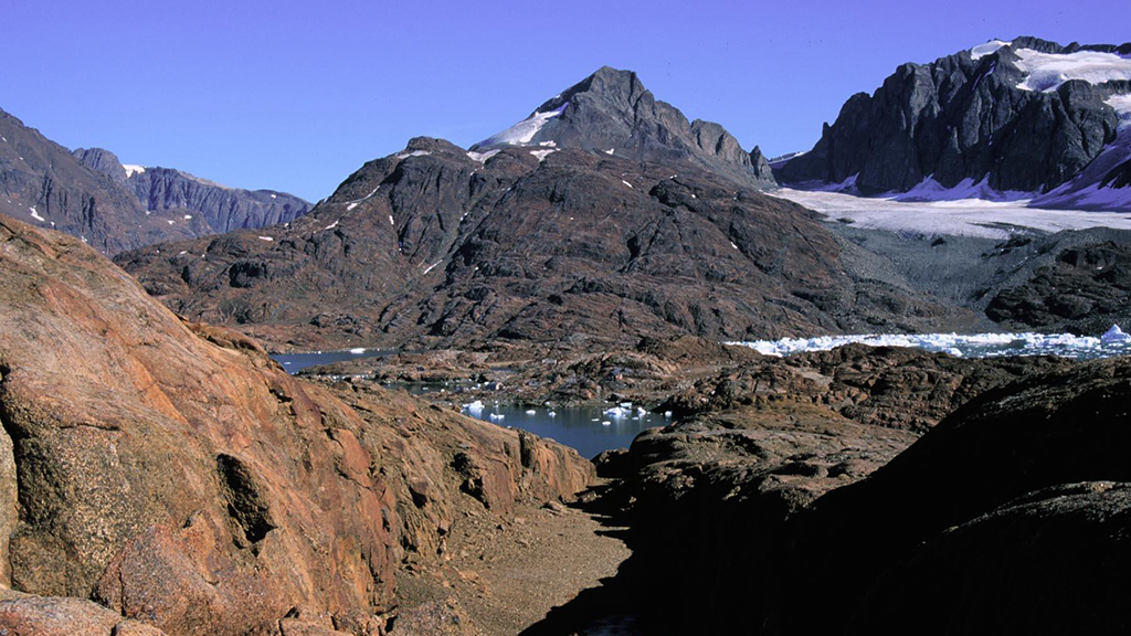 Mountains, rocks in the foreground, a glacier between mountains, and water in a valley.