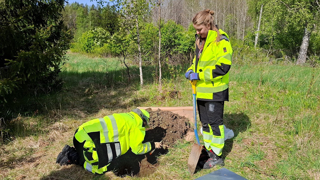 Kaksi henkilöä kirkkaissa huomiovaatteissa tekee maastotutkimusta: toinen kaivaa kuoppaa ja toinen seisoo lapion kanssa metsäisellä alueella.