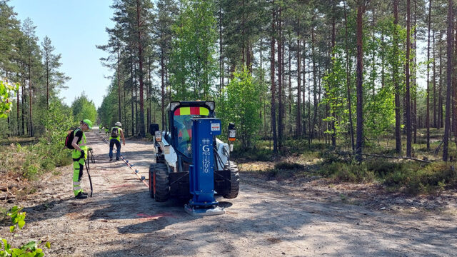Kaksi mittaajaa tekee maastomittauksia metsätiellä, ja heidän vieressään on mittauslaite pientraktorin edessä.