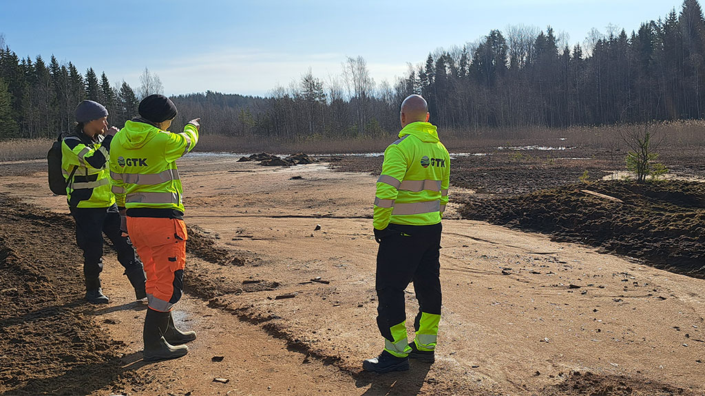 Three people stand on a tailings area. One person points at the terrain. The people wear high-visibility clothing.