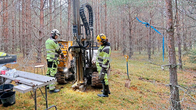 Kaksi henkilöä käyttää kairauslaitetta metsässä. Laitteen vieressä on pöytä, työvälineitä ja lapio. Puita ympärillä.