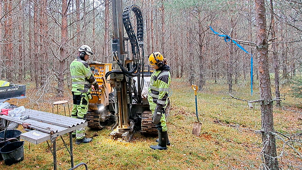 Två personer använder en borrigg i en skog. Ett bord, verktyg och en spade finns bredvid utrustningen. Träd runt omkring.