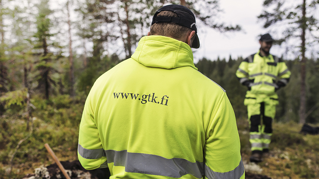 Kaksi maastotyöntekijää keltaisissa suojavaatteissa metsässä; etualalla olevan selässä teksti “www.gtk.fi”.