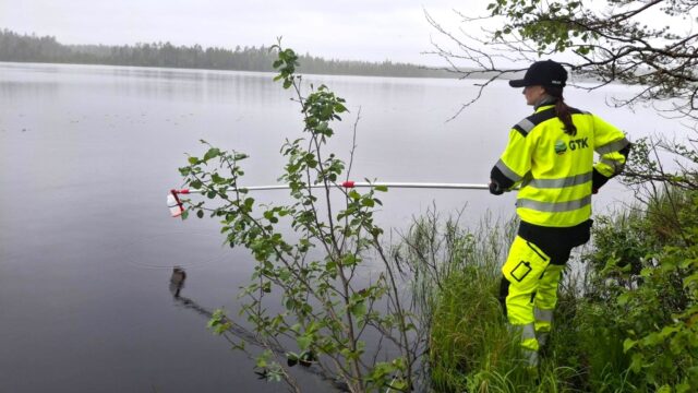 Henkilö keltaisissa huomiovaatteissa haavin kanssa järven äärellä.