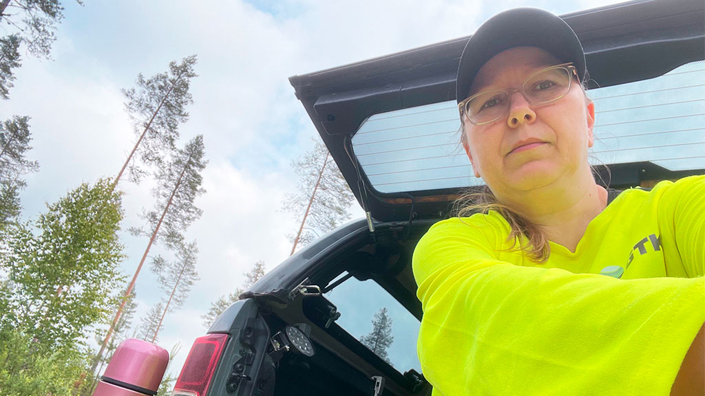 Henkilö istuu auton tavaratilassa. Hänellä on termospullo. Taustalla metsämaisema ja puita.