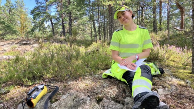 Henkilö istuu kalliolla keltaisissa huomiovaatteissa ja hymyilee kameralle.