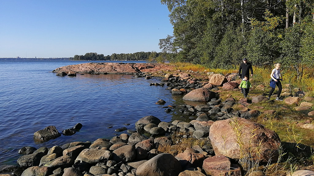 Maisema suomalaisesta merenrannasta kesällä. Kolme henkilöä kävelee rantakivikossa rannan tuntumassa. Vasemmalla näkyy meri ja rannassa kasvaa vihreää lehtimetsää, pensaita ja kaislikkoa.