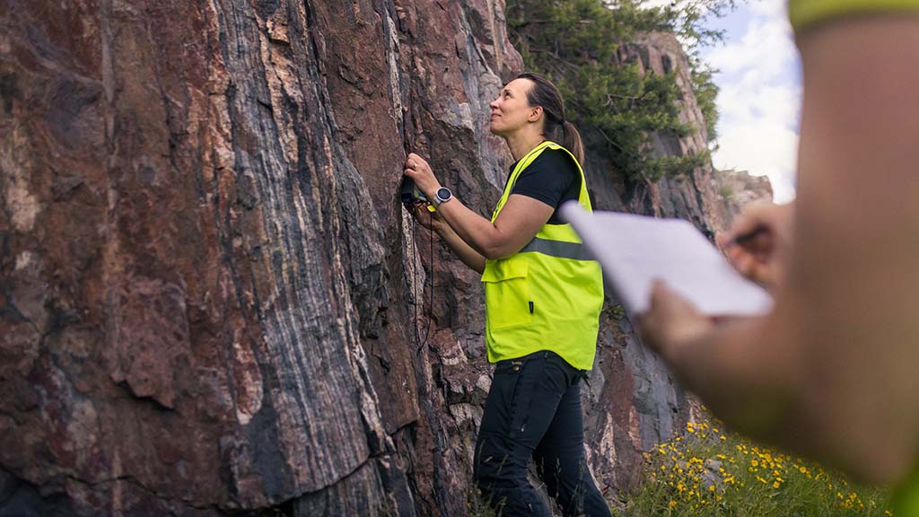 Henkilö huomioliivissä tutkii kallioseinämää, toinen henkilö tekee muistiinpanoja taustalla.