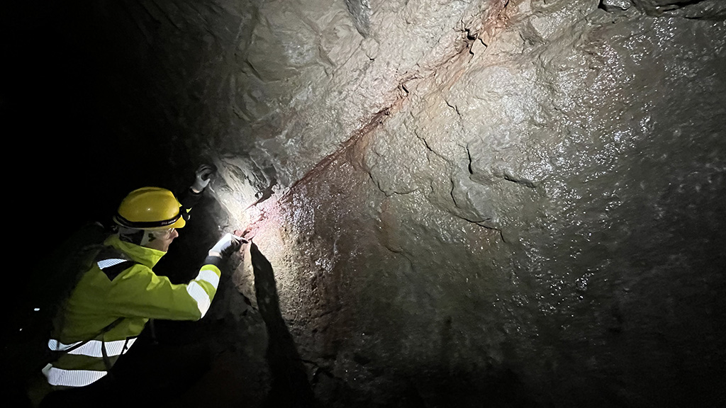 Suojakypärään ja -vaatteisiin pukeutunut henkilö tutkii kallioseinämän rakoilua tunnelissa valon avulla.