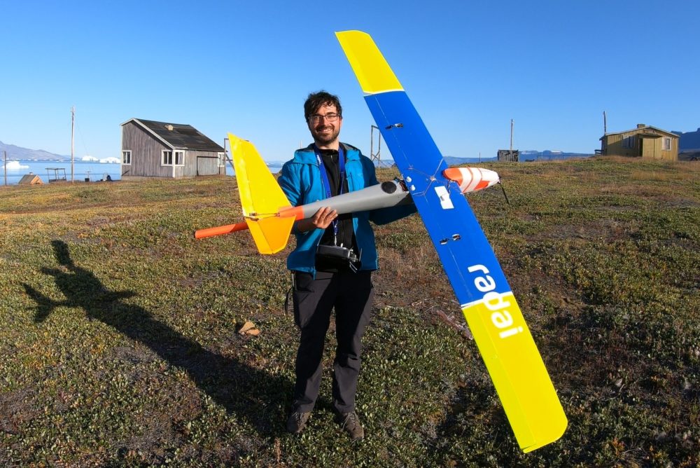 Figure 2. Radai Oy’s fixed-wing drone equipped with a fluxgate magnetometer (orange cylinder) in the tail boom. Robert Jackisch (HZDR-HIF) is holding the drone. The photo is from a field campaign in Greenland in 2019 (Photo: Heikki Salmirinne, GTK).