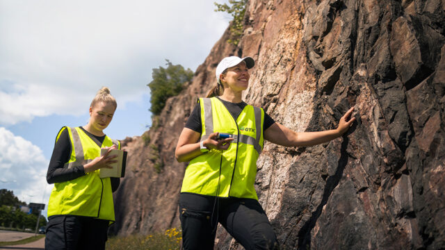 Kesäinen poutapilvinen maisema, jossa kaksi Geologian tutkimuskeskuksen naistutkijaa katsastavat kallioseinämää.