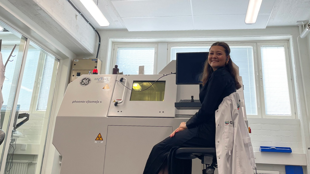 Hymyilevä henkilö istuu tutkimuslaboratoriossa röntgentomografialaitteen äärellä ja katsoo kameraan