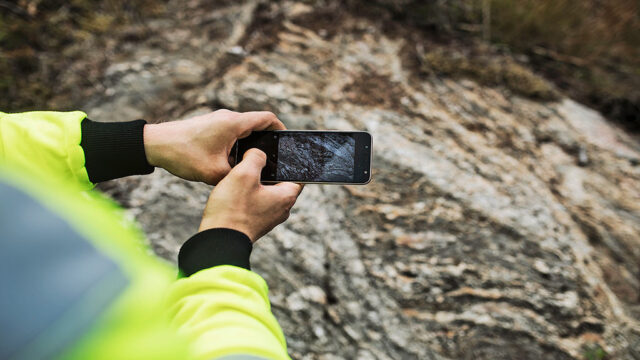 Lähikuva mobiililaitteesta ja käsistä. Otetaan kuvaa kalliosta.