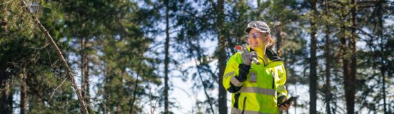 Nainen huomiovaatteissa metsässä hakku olalla.