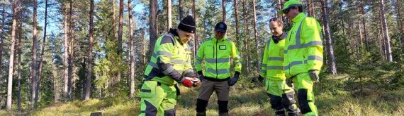 Neljä miestä tarkkailevat maassa olevaa mittalaitetta mäntymetsässä.