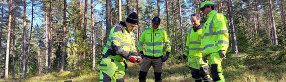 Neljä miestä tarkkailevat maassa olevaa mittalaitetta mäntymetsässä.